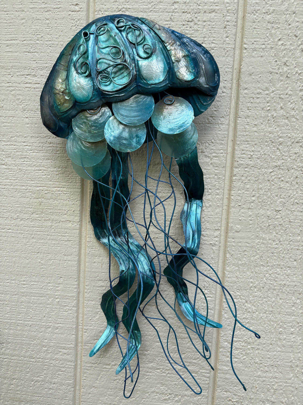 May include: A blue and green metal jellyfish wall sculpture with a textured, iridescent finish and capiz shells. The jellyfish has a roundish head with a swirling design and a long, flowing tail made of wire.